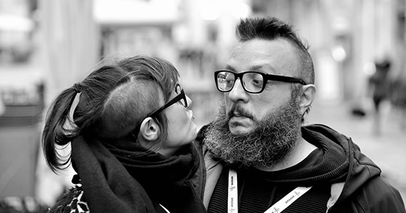 a woman and a man look into each other's eyes. he has a beard and wears a hoody; she has long hair gathered in a ponytail with shaved sides