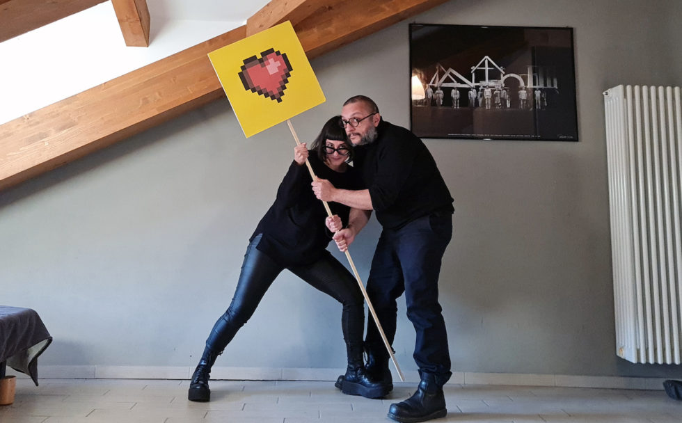 A woman and a man pose in a room with low ceiling. they hold a sign with a pixelated heart